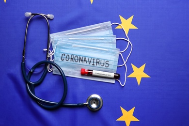 Photo of Stethoscope, protective masks and test tube with blood sample on European Union flag background, flat lay. Coronavirus outbreak