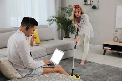 Photo of Man working on laptop while his wife cleaning carpet in living room. Stay at home concept