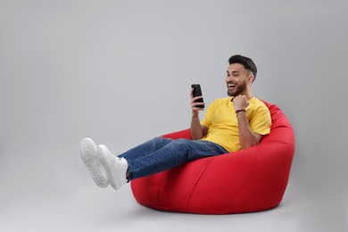 Happy young man using smartphone on bean bag chair against grey background, space for text