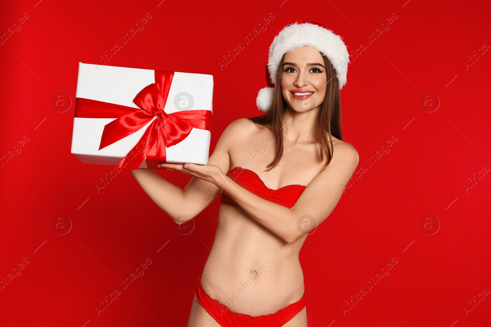 Photo of Happy young woman in bikini and Santa hat with gift box on red background. Christmas celebration
