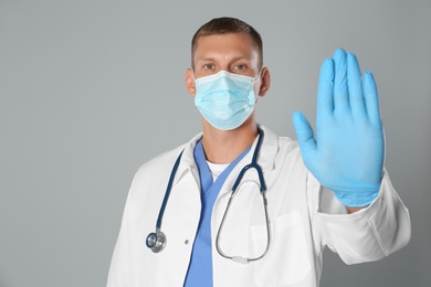 Photo of Doctor in protective mask showing stop gesture on grey background. Prevent spreading of coronavirus