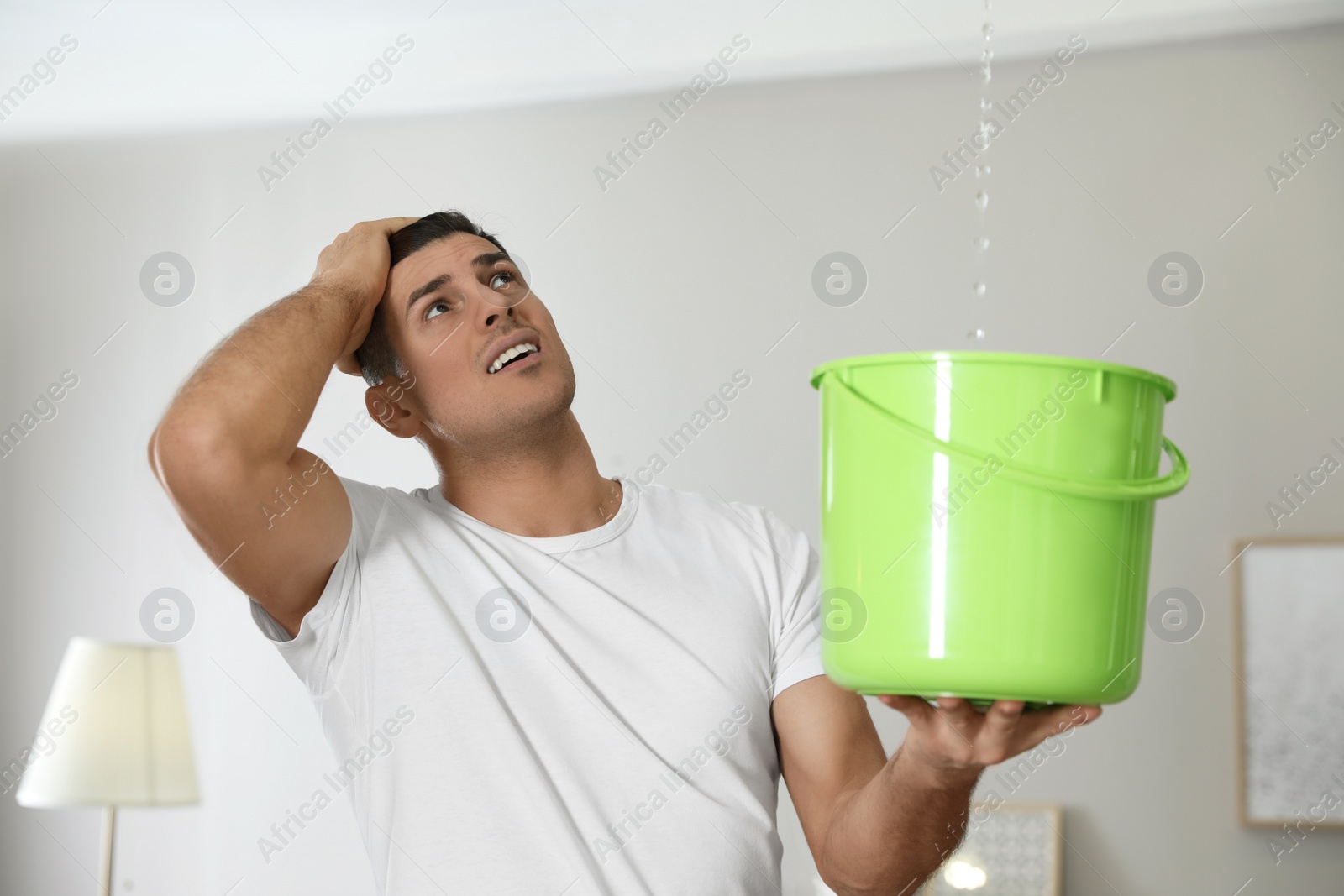 Photo of Emotional man collecting water leaking from ceiling in living room. Damaged roof