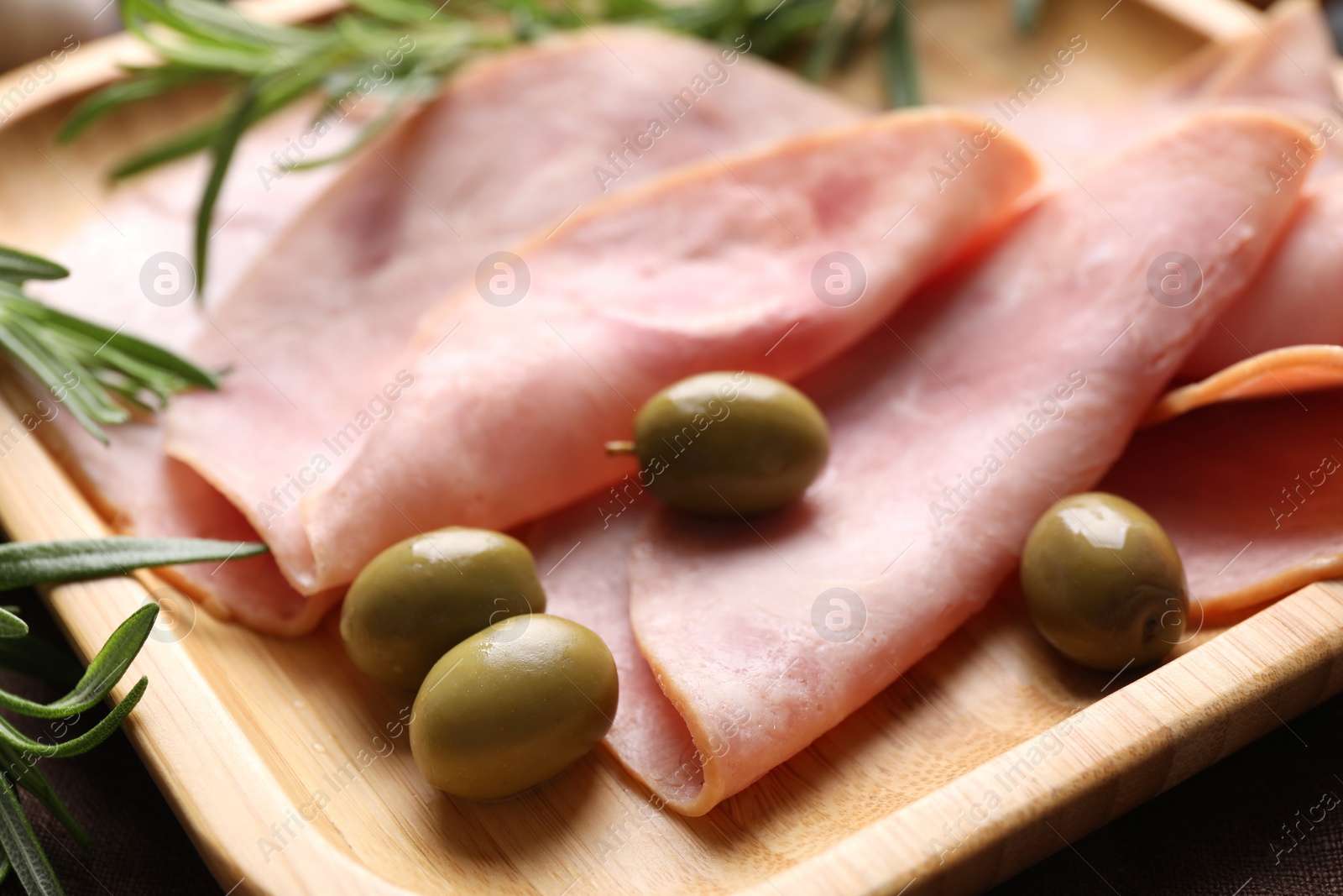 Photo of Slices of delicious ham with rosemary and olives on wooden tray, closeup