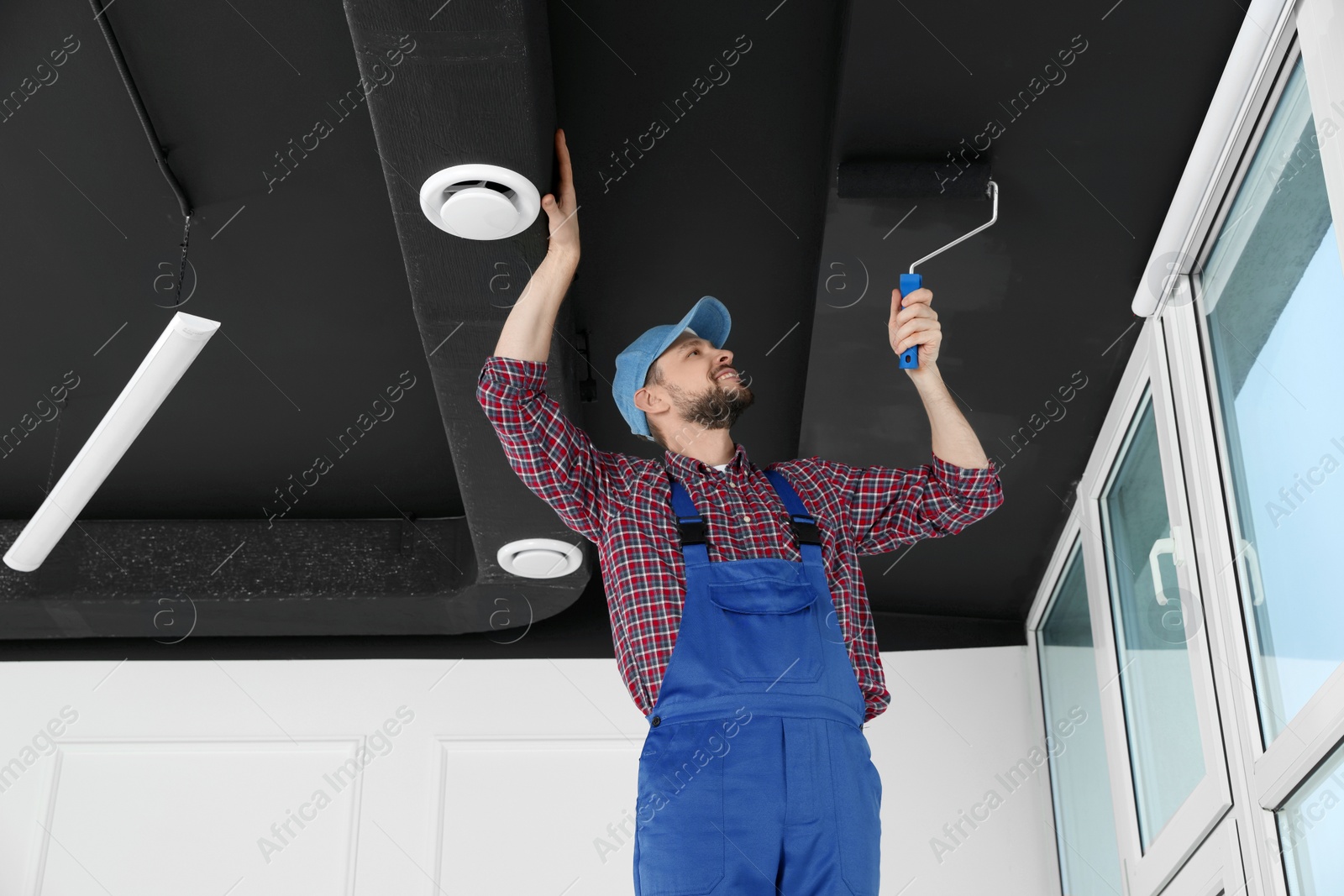 Photo of Worker in uniform painting ceiling with roller indoors, low angle view