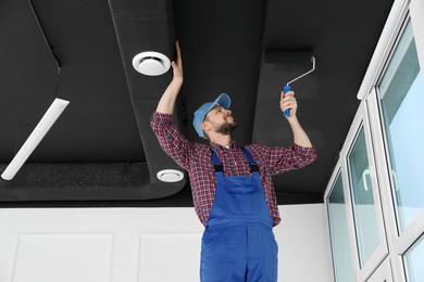 Photo of Worker in uniform painting ceiling with roller indoors, low angle view