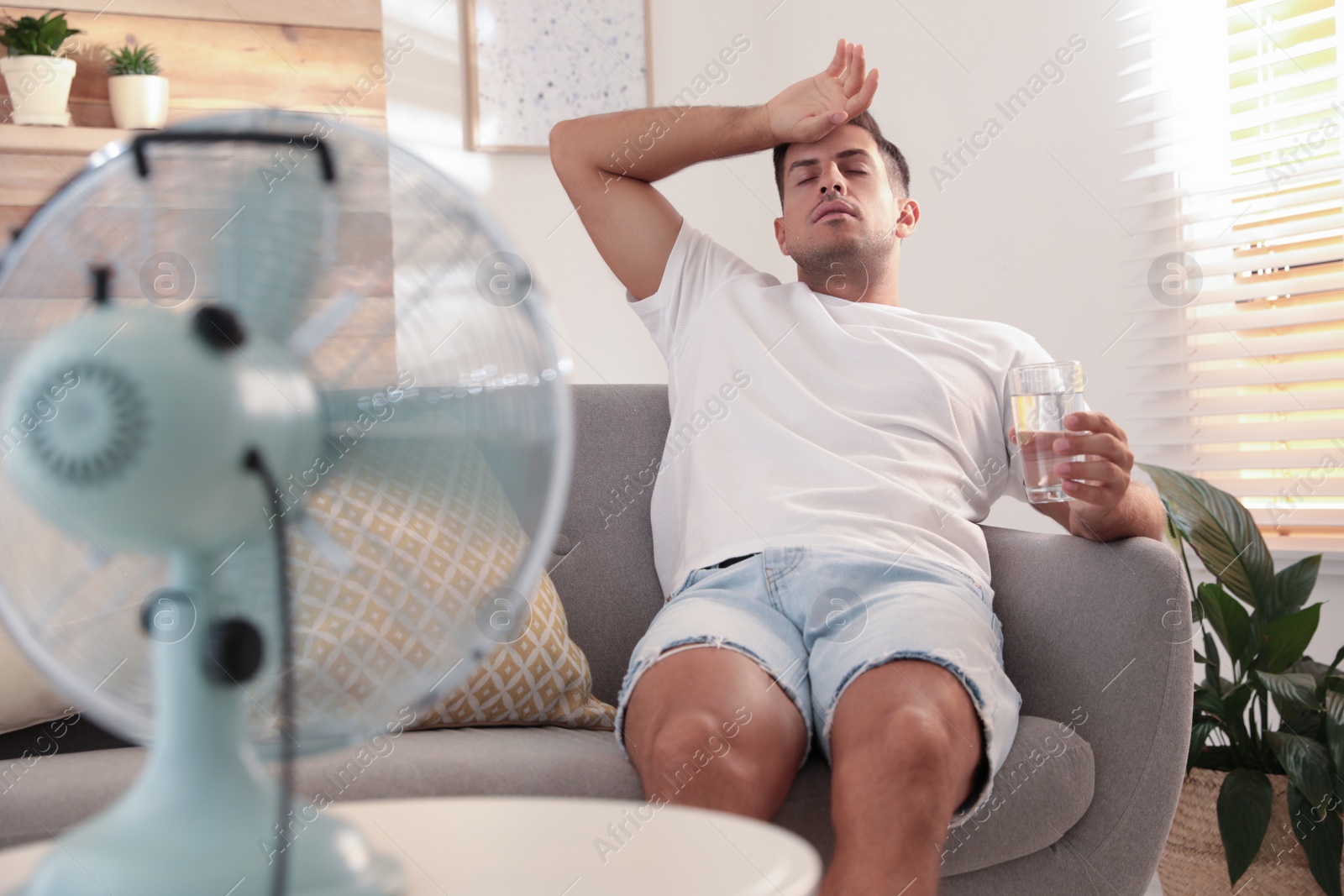 Photo of Man with water sitting on sofa near modern fan in living room. Summer heat