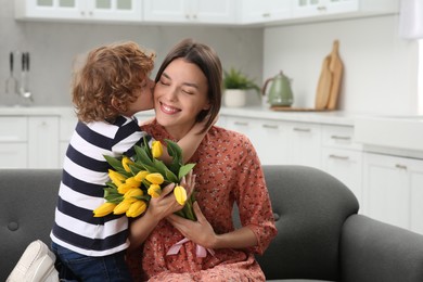 Little son kissing and congratulating his mom with Mother`s day at home. Woman holding bouquet of yellow tulips