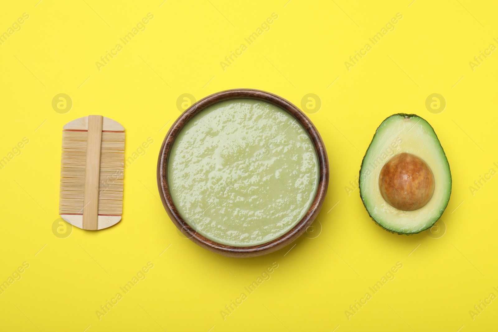 Photo of Homemade hair mask in bowl, avocado and bamboo comb on yellow background, flat lay