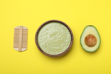 Homemade hair mask in bowl, avocado and bamboo comb on yellow background, flat lay