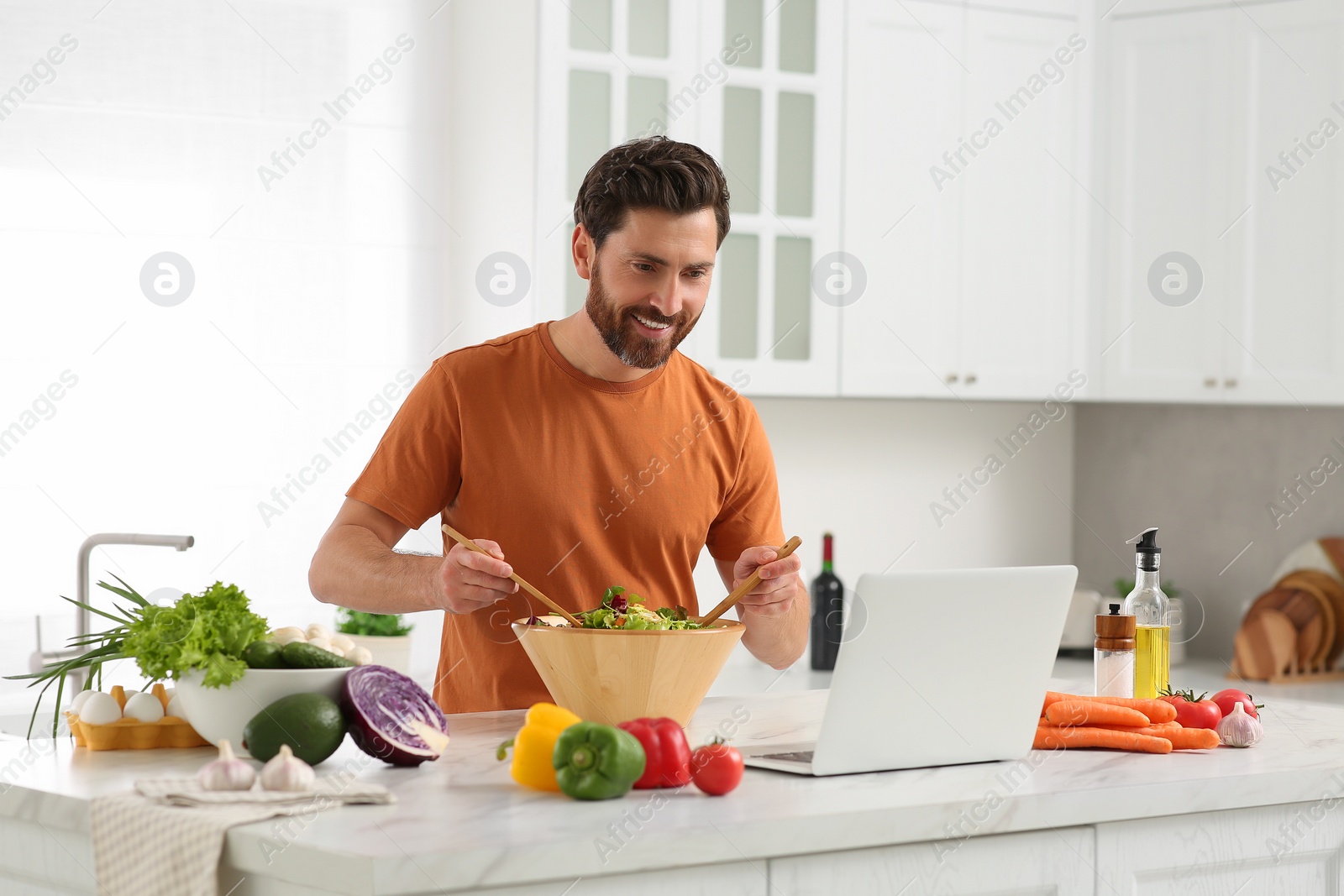 Photo of Man making dinner while watching online cooking course via laptop in kitchen