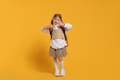 Photo of Happy schoolgirl with backpack showing thumbs up gesture on orange background