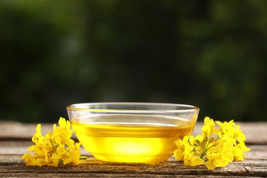 Photo of Rapeseed oil in glass bowl and beautiful yellow flowers on wooden table outdoors