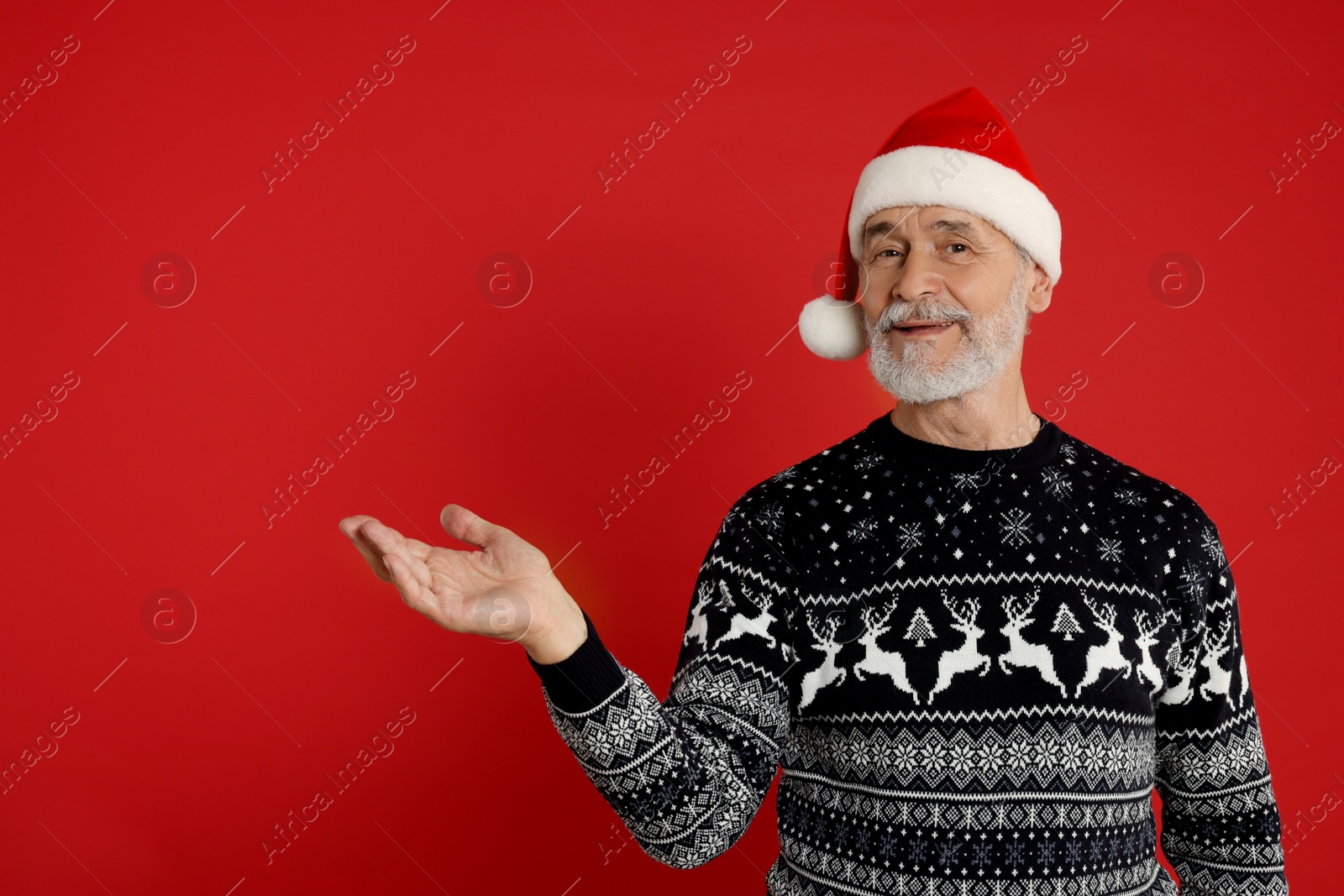 Photo of Senior man in Christmas sweater and Santa hat showing something on red background