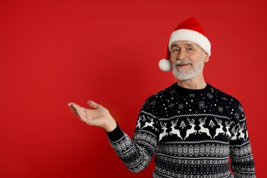 Senior man in Christmas sweater and Santa hat showing something on red background