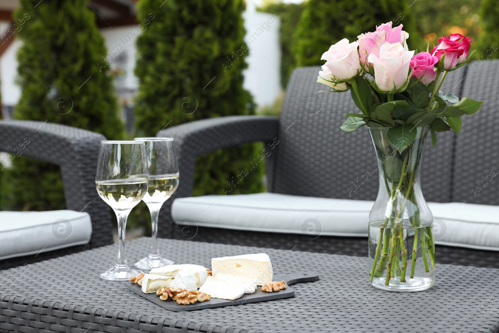 Photo of Vase with roses, glasses of wine and food on rattan table on outdoor terrace