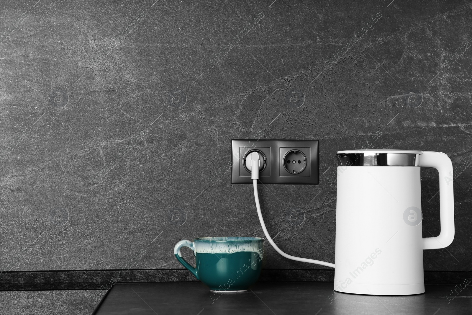 Photo of Electric kettle plugged into power socket on dark grey wall indoors. Space for text