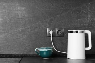 Electric kettle plugged into power socket on dark grey wall indoors. Space for text