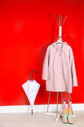 Photo of Stylish umbrella, raincoat and boots near red wall