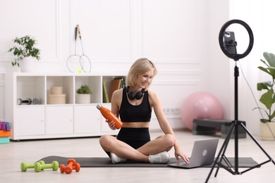 Photo of Smiling sports blogger holding bottle while streaming online fitness lesson at home