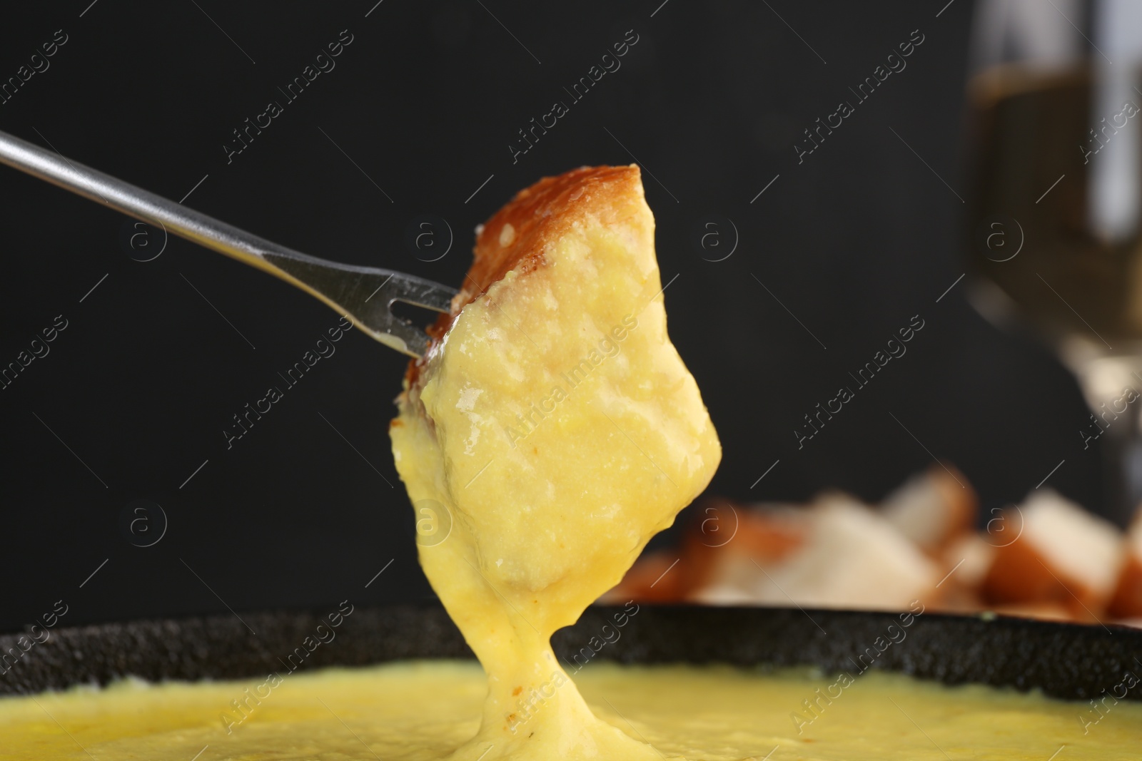 Photo of Dipping piece of bread into fondue pot with tasty melted cheese against dark gray background, closeup