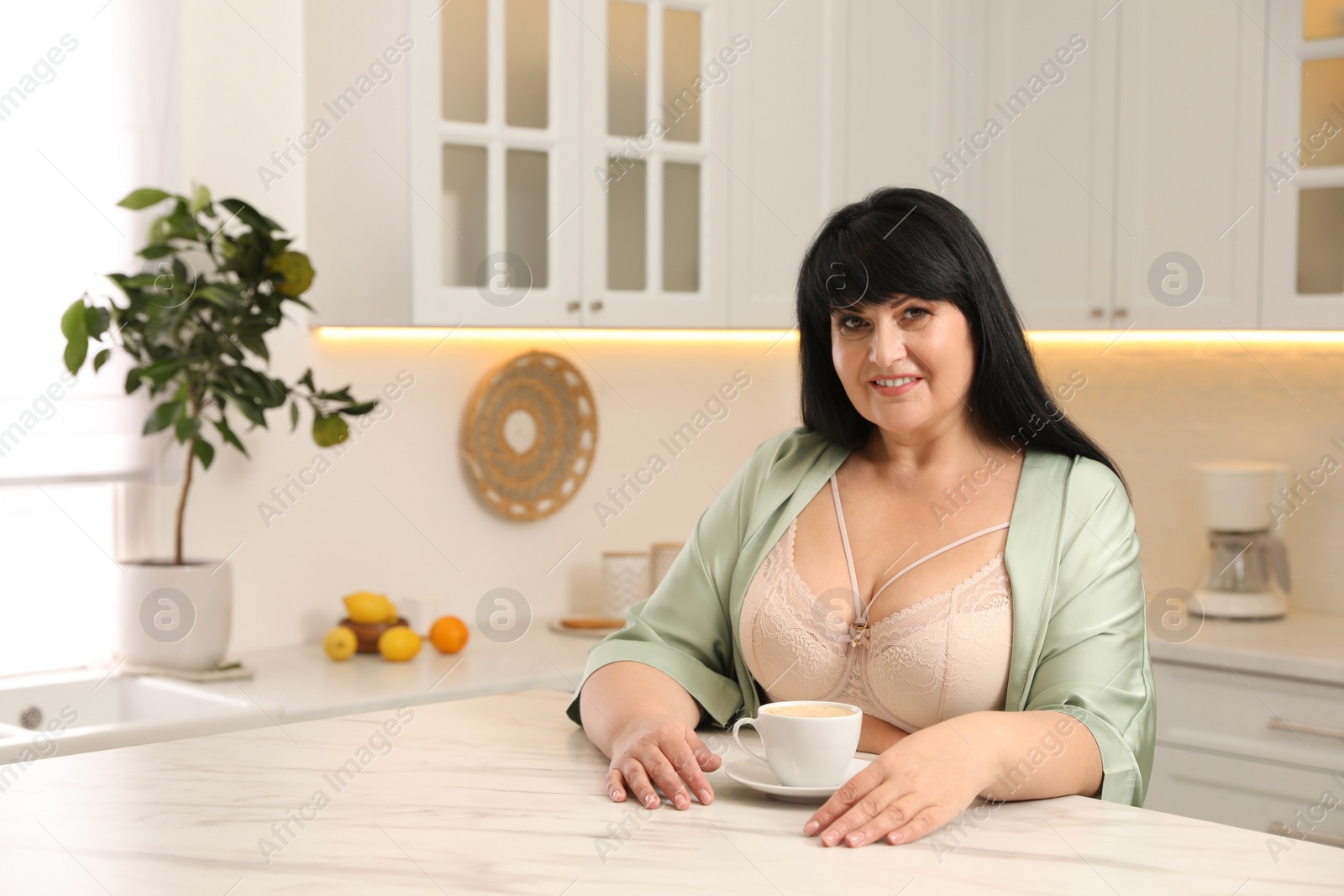 Photo of Beautiful overweight woman in beige underwear and silk robe with cup of coffee at kitchen table. Plus-size model