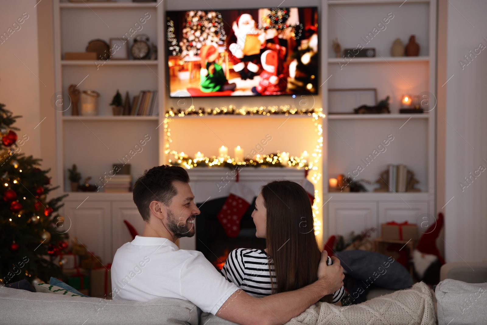 Photo of Happy couple spending time near TV in cosy room, back view. Christmas atmosphere