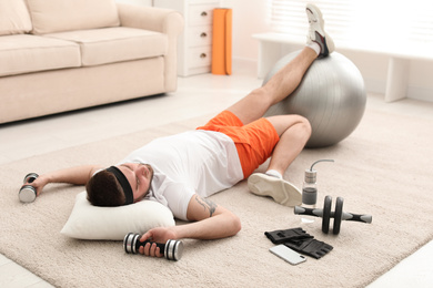 Photo of Lazy young man with sport equipment sleeping on floor at home
