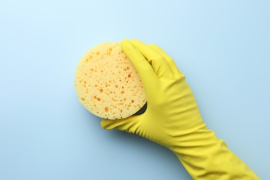 Photo of Cleaner in rubber glove holding new yellow sponge on light blue background, top view.