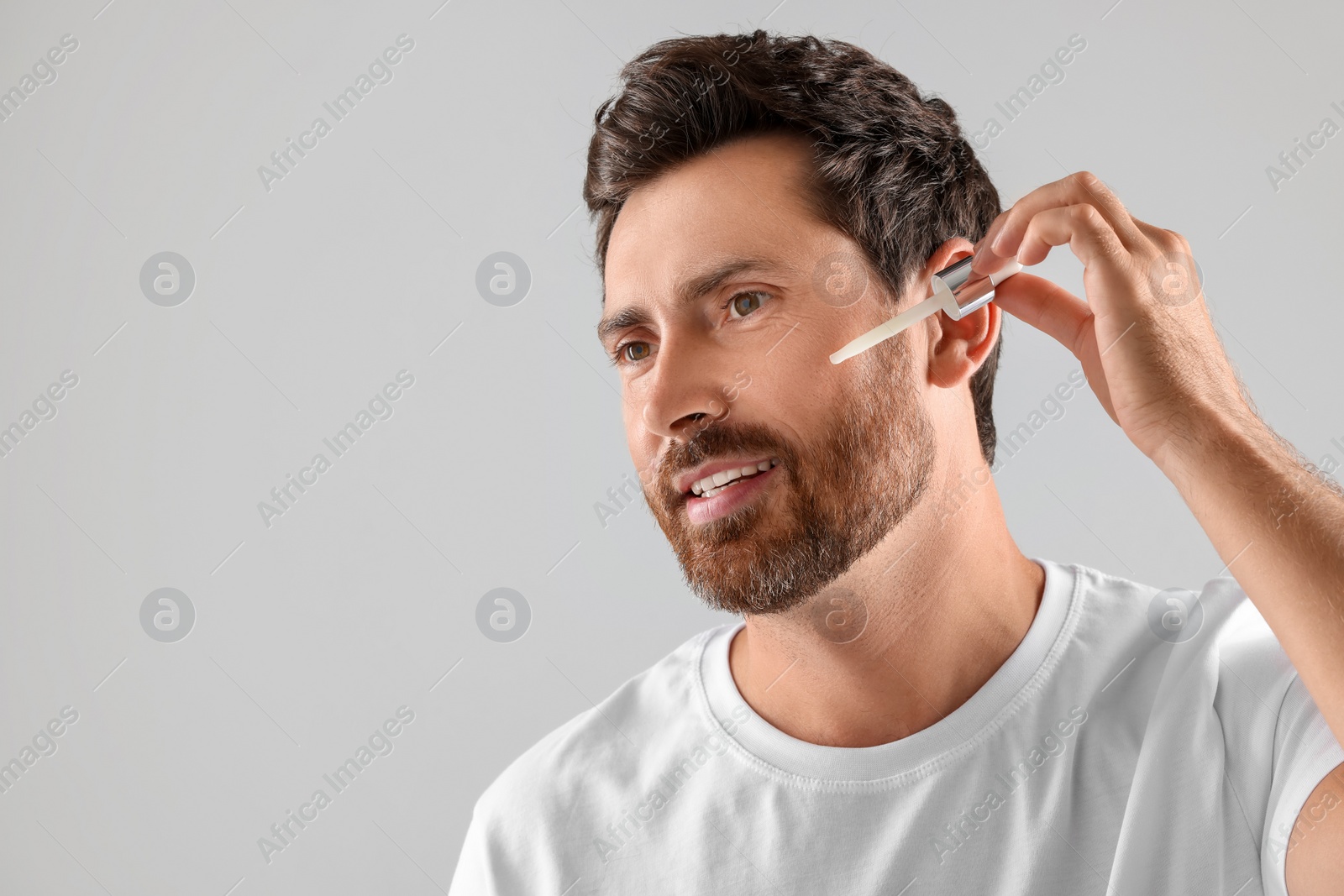 Photo of Smiling man applying cosmetic serum onto his face on light grey background. Space for text