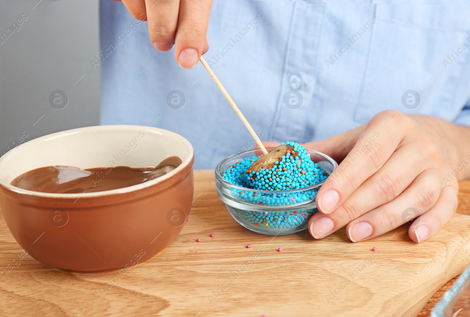 Photo of Young woman putting cake pop into blue sprinkles on wooden board, closeup