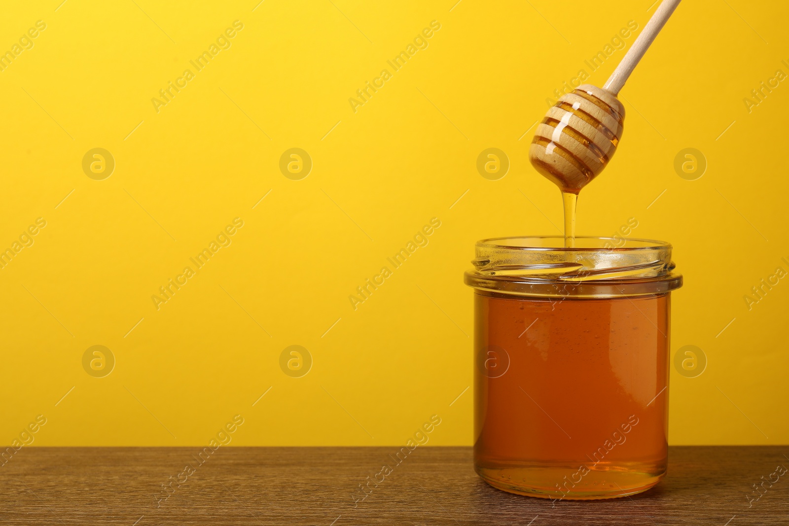 Photo of Pouring honey from dipper into jar at wooden table against yellow background, space for text