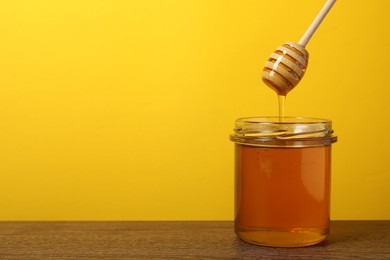 Pouring honey from dipper into jar at wooden table against yellow background, space for text