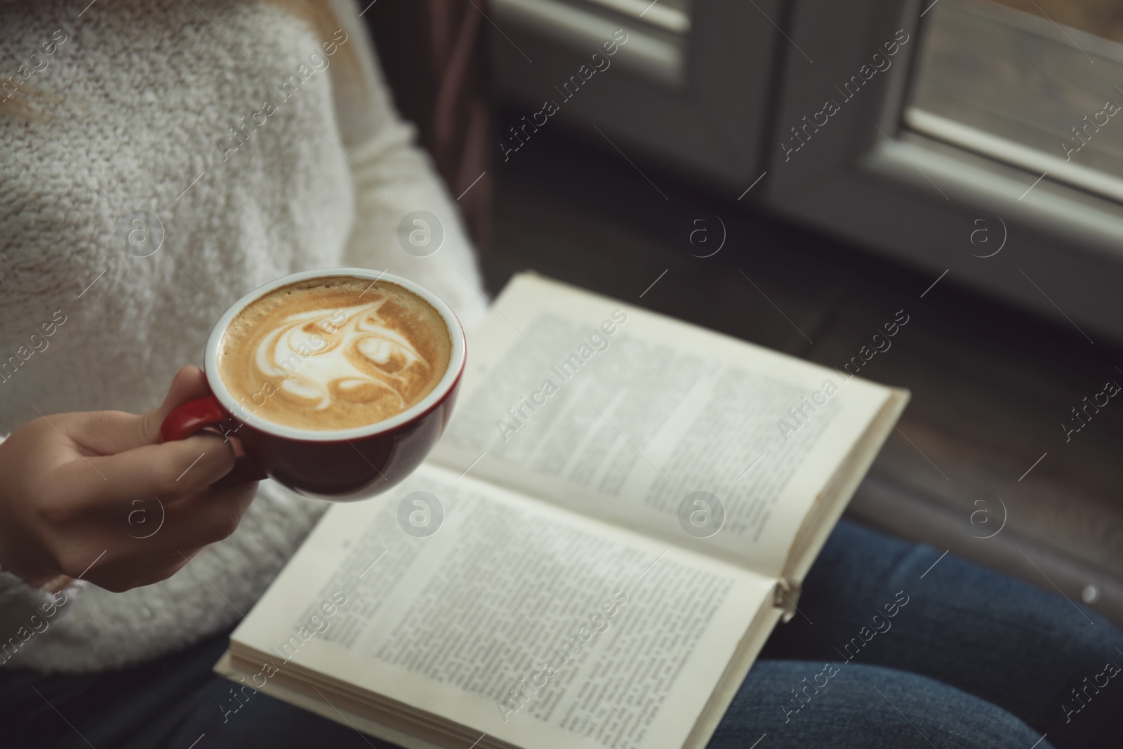 Photo of Woman with cup of coffee reading book near window indoors, closeup