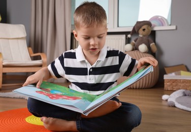 Photo of Cute little boy reading book on floor at home