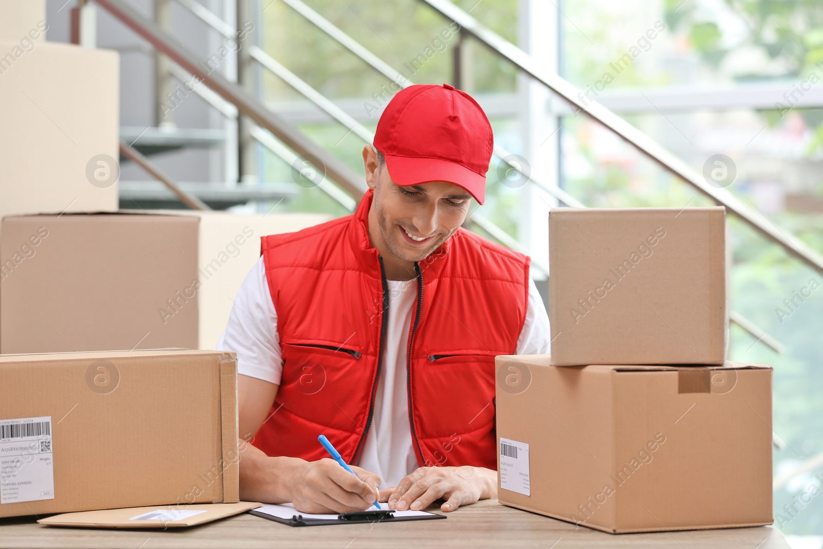 Photo of Young courier working with papers among parcels at table in delivery department