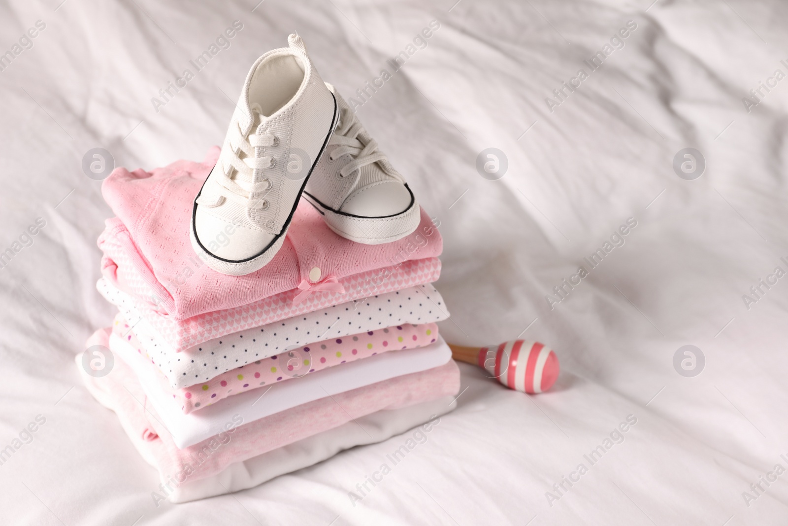 Photo of Stack of baby girl's clothes, rattle and shoes on bed. Space for text