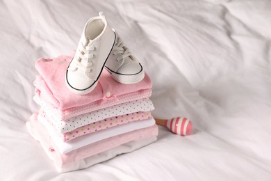 Stack of baby girl's clothes, rattle and shoes on bed. Space for text