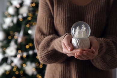 Photo of Woman in sweater holding decorative snow globe against Christmas tree, closeup. Space for text