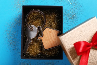 Photo of Key with trinket in shape of house, gift box and glitter on light blue background, flat lay. Housewarming party