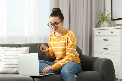 Home workplace. Happy woman with smartphone and laptop on sofa in room