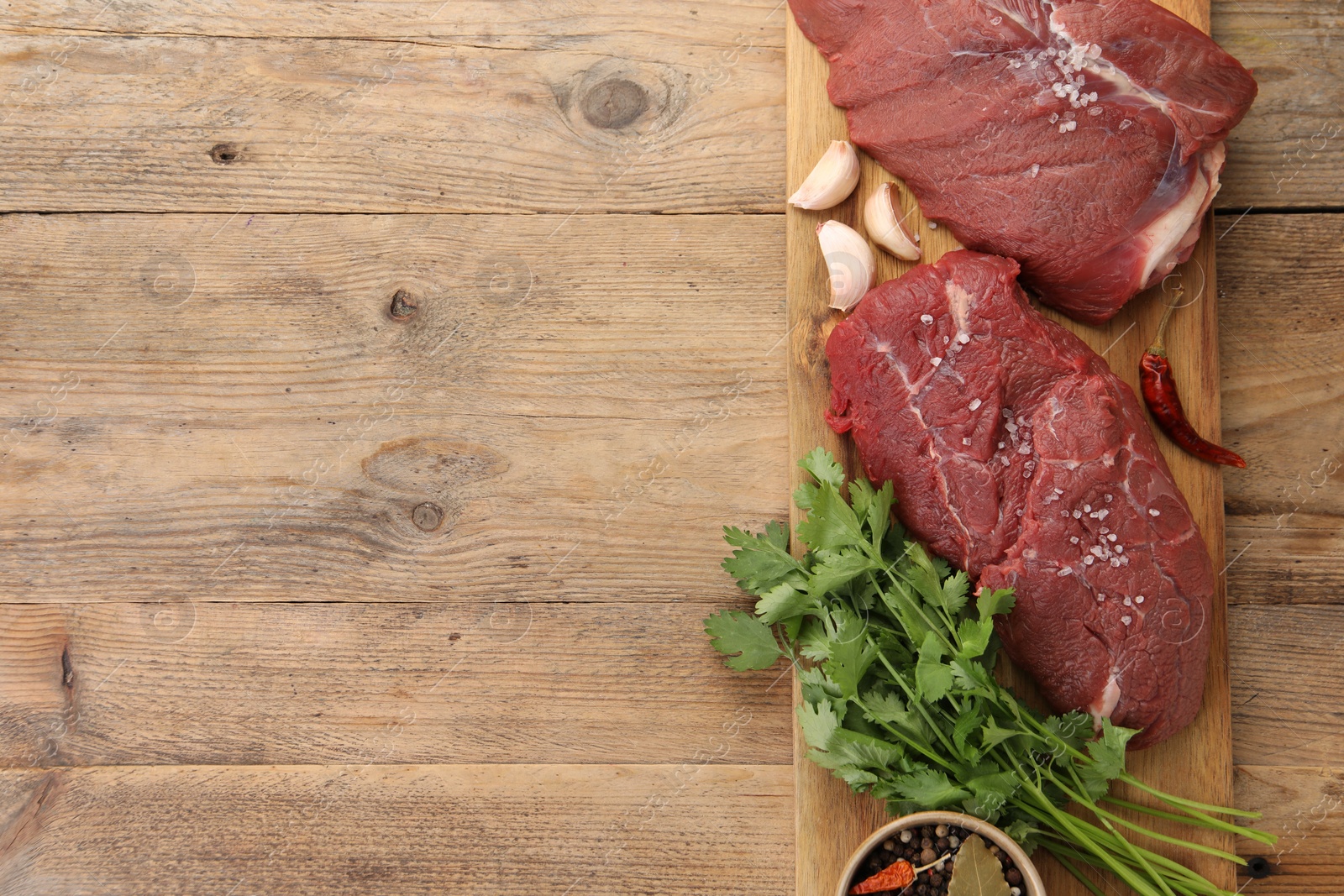 Photo of Pieces of raw beef meat, parsley, garlic and spices on wooden table, top view. Space for text