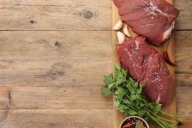 Photo of Pieces of raw beef meat, parsley, garlic and spices on wooden table, top view. Space for text