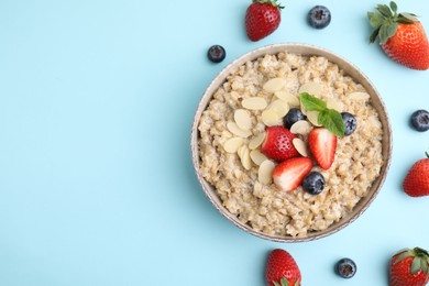 Photo of Tasty oatmeal with strawberries, blueberries and almond flakes in bowl surrounded by fresh berries on light blue background, flat lay. Space for text