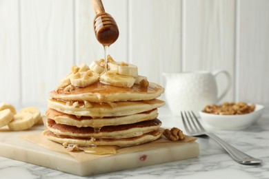 Photo of Pouring honey from dipper onto delicious pancakes with bananas and walnuts at white marble table, closeup