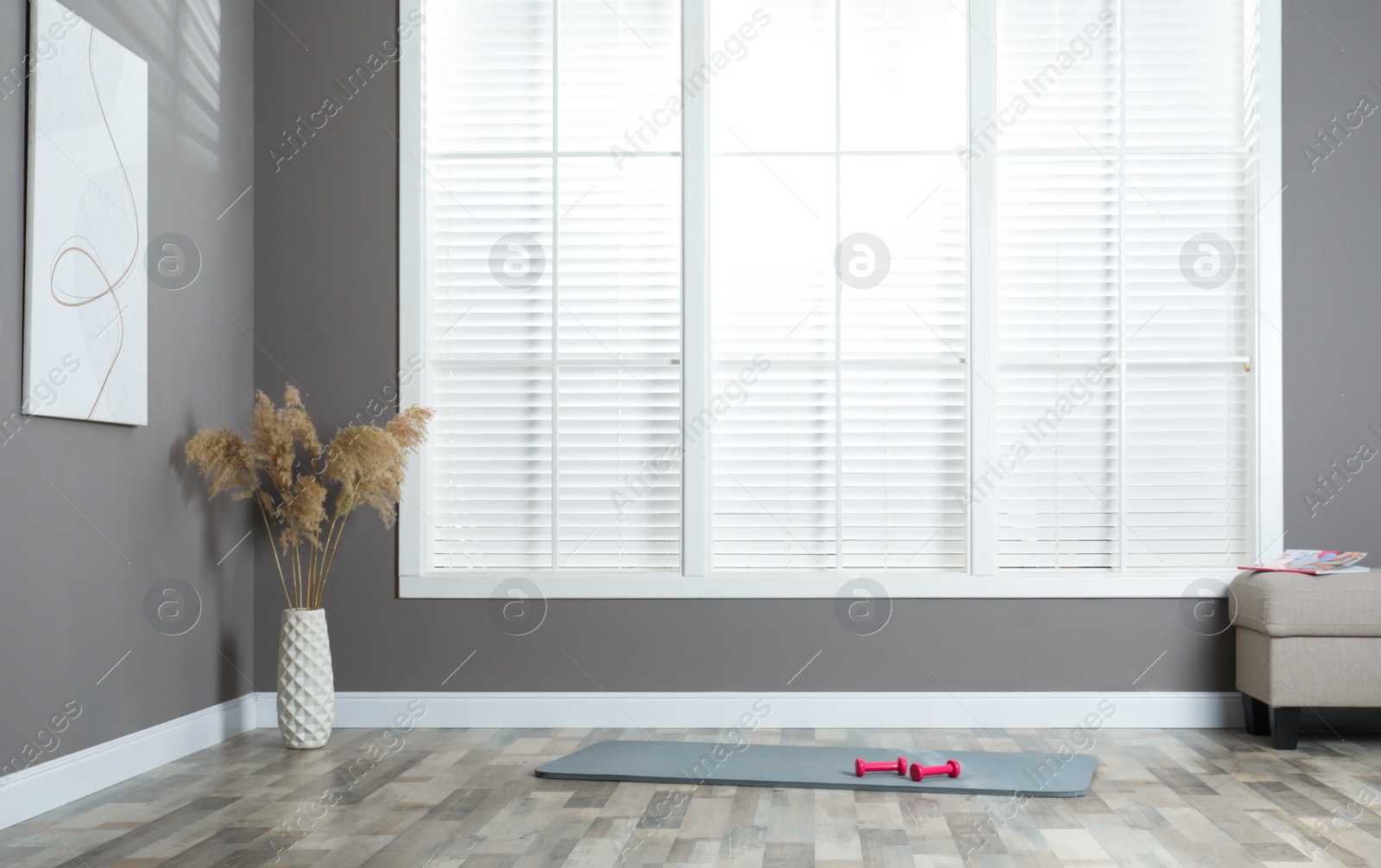 Photo of Exercise mat with dumbbells near vase of dried reeds and pouf in spacious room