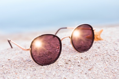 Stylish sunglasses and starfish on sandy beach near sea, closeup