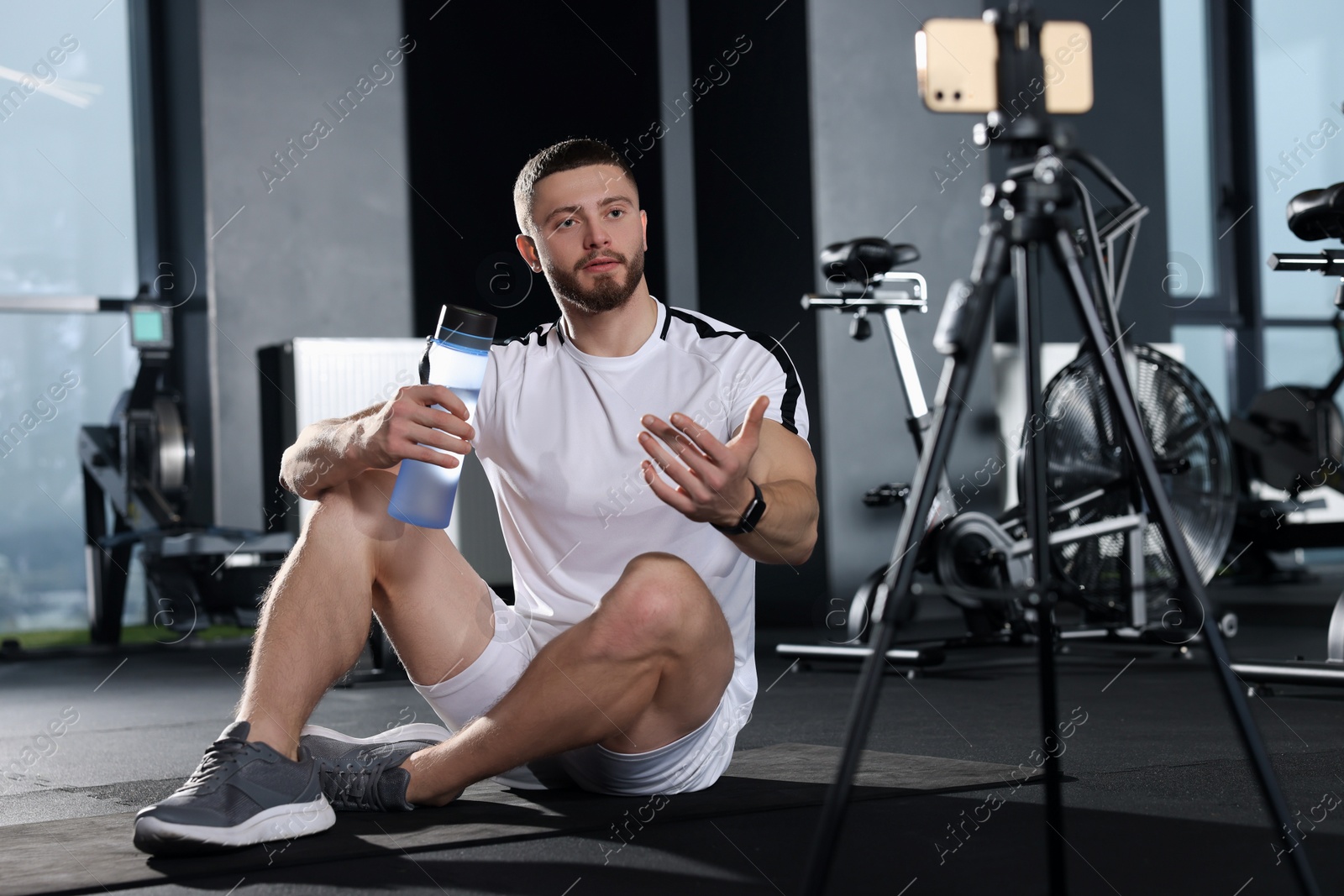 Photo of Man with bottle of water streaming online training on phone at gym. Fitness coach
