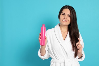 Photo of Beautiful young woman in bathrobe holding bottle of shampoo on light blue background, space for text