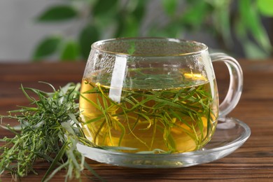 Photo of Cup of homemade herbal tea and fresh tarragon leaves on wooden table, closeup