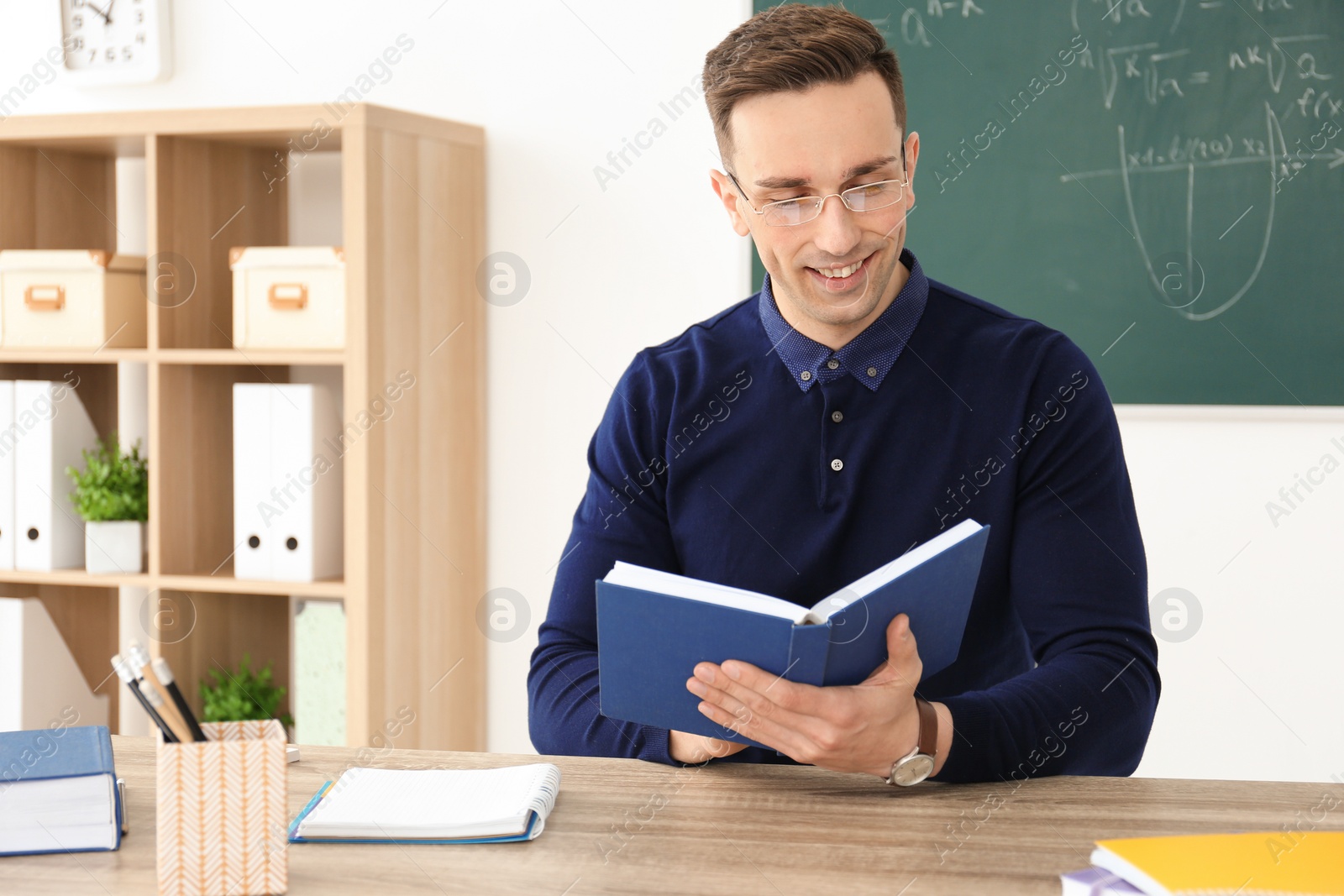 Photo of Young male teacher with book sitting at table in classroom